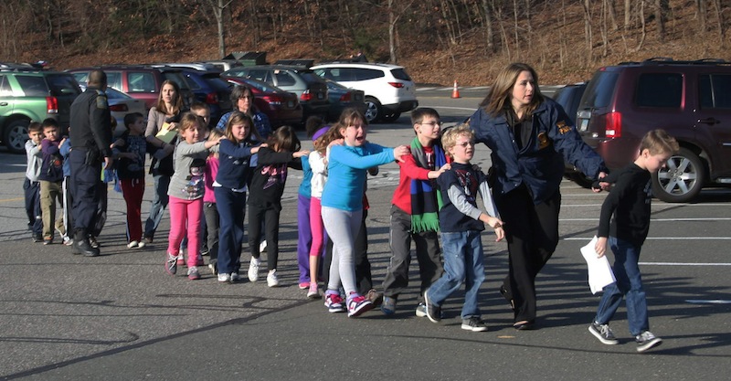 In this Friday, Dec. 14, 2012 file photo provided by the Newtown Bee, Connecticut State Police lead a line of children from the Sandy Hook Elementary School in Newtown, Conn. after a shooting at the school. The private equity firm Cerberus will sell its stake in a firearms company that produced one of the weapons believed to have been used in the shootings at the elementary school, calling it a "watershed event" in the national debate on gun control. While saying that it's not its role to take positions or attempt to shape or influence the gun control debate, Cerberus said it is taking what action it can by selling its stake in the Freedom Group, which makes the Bushmaster rifle. (AP Photo/Newtown Bee, Shannon Hicks)