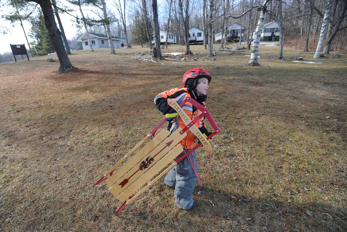 Mario Wilbur, 7, of East Madison, lugs his Flexible Flyer across the grassy hill en route to the ice fishing derby at Lake George, on the Skowhegan and Canaan town line, on Saturday. Wilbur said that even though there is no snow, the sled works perfectly fine on the frozen lake.