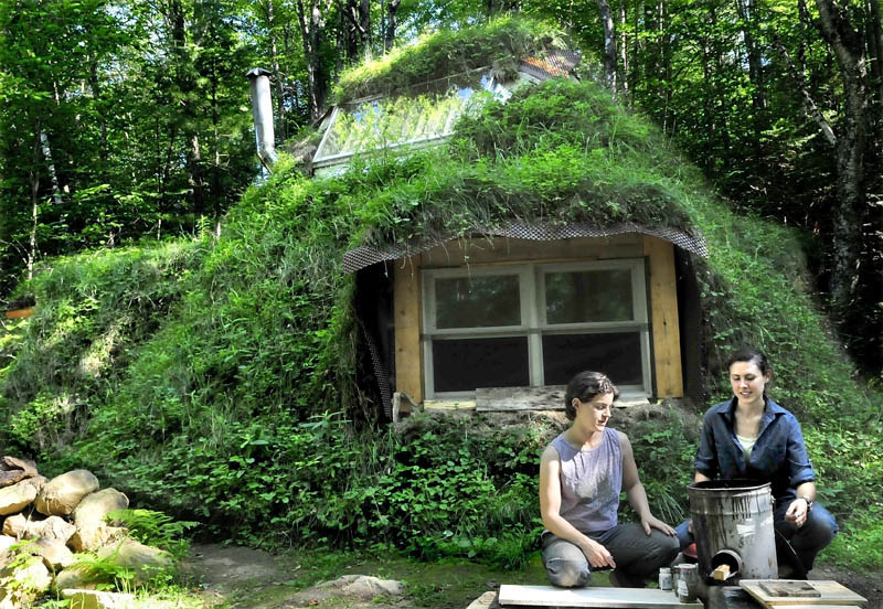 Ashirah Knapp, left, and apprentice Ashley Hardy look over the highly efficient wood stove that Hardy used to boil water earlier in the day at the Koviashuvik Living School in Starks recently. Behind the women is the wood-and-soil-covered apprentice lodge where Hardy stayed.