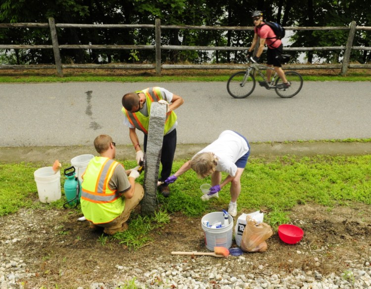 A passing cyclist watches city workers Taylor Perry, left, and Shawn Winslow and volunteer Kathleen “Sukey” Sikora remove graffiti from a Kennebec River Rail Trail milestone on Wednesday in Augusta.
