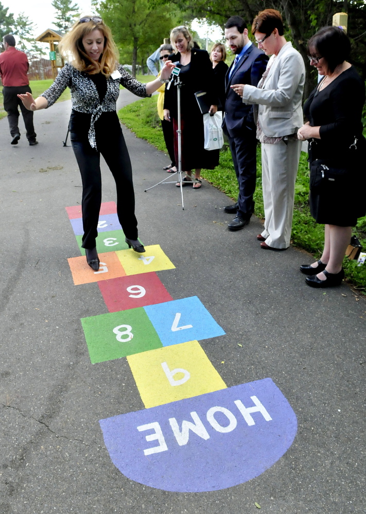Tina Chapman of the Waterville Rotary Club is cheered on as she demonstrates hopscotch, one of 10 stations along a walking trail in Waterville, during a press conference on Thursday to bring attention to the trail system and encourage parents and children to use.