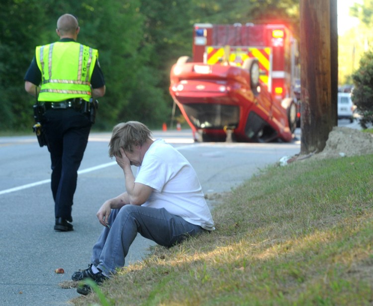 James Salisbury, foreground, holds his head in his hand as Waterville police Officer Chase Fabian walks towards the wreckage of Salisbury’s car on upper Main Street in Waterville on Wednesday. Salisbury hit the power pole, flipping his car on to its roof.