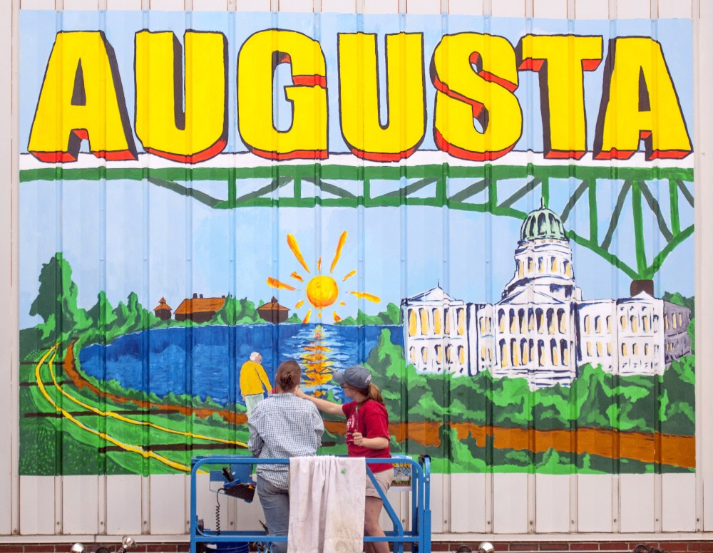 Staff photo by Joe Phelan
University of Maine at Augusta art students Marcea Crawford, left, and Sierra Meservey work on June 15 in a scissor lift on a large postcard-style mural on the side of 179 Water St. overlooking the intersection of Bridge and Water streets in downtown Augusta. It was the first downtown mural to be completed, with others in the works.