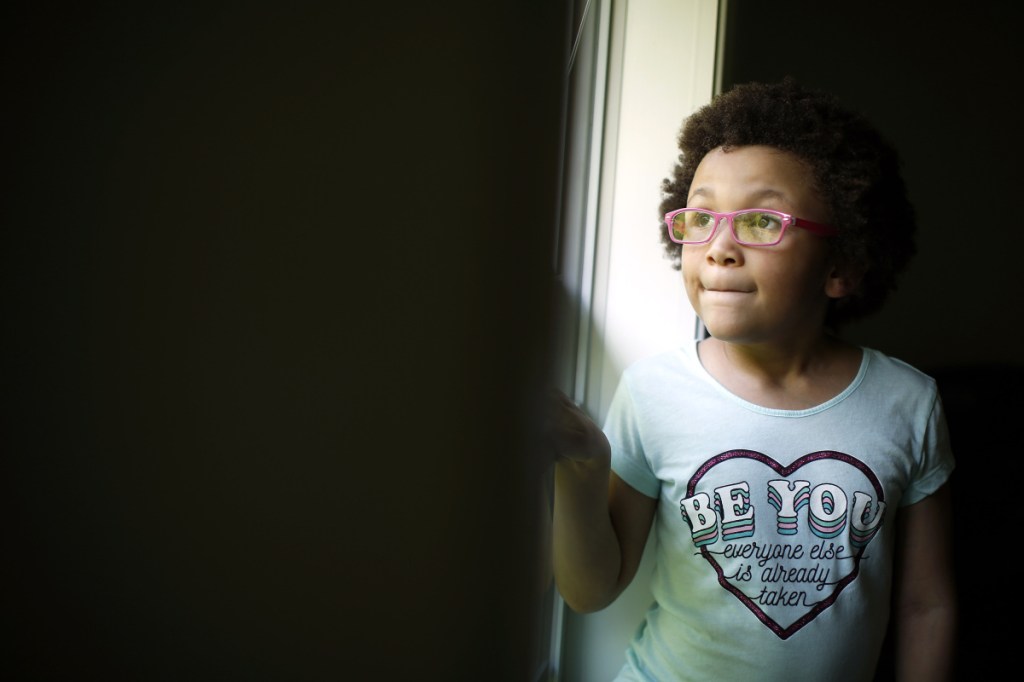 Zoé Paul, 6, looks at some neighborhood children playing outside her bedroom window in Windham. For now, Zoé, who has a weakened immune system, is being home-schooled because her parents are concerned about their daughter being around unvaccinated students.