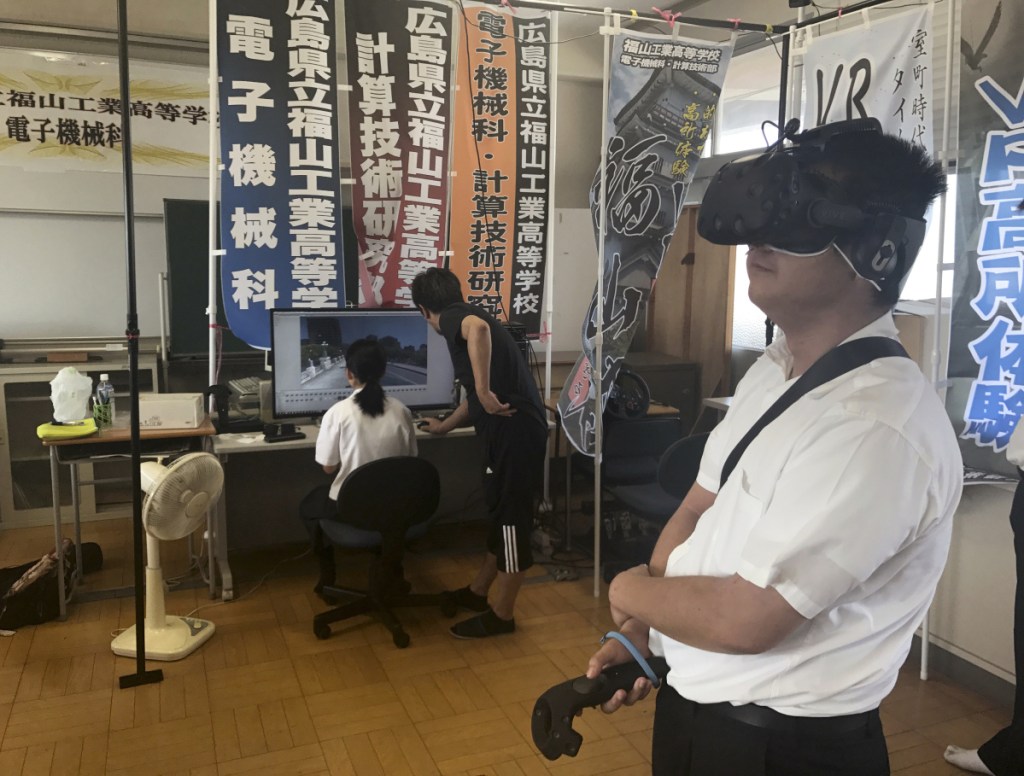 In Friday, Aug. 8, 2018, photo, Namio Matsura, right, a 17-year-old member of the computation skill research club at Fukuyama Technical High School, watches Hiroshima city before atomic bomb fell in virtual reality experience as Katsushi Hasegawa, a computer teacher, and Mei Okada, another member of the club, watch a computer screen, at the school in Hiroshima, western Japan. Over two years, the group of Japanese high school students has been painstakingly producing a five-minute virtual reality experience that recreates the sights and sounds of Hiroshima before, during and after the U.S. dropped an atomic bomb on the city 73 years ago. (AP Photo/Haruka Nuga)