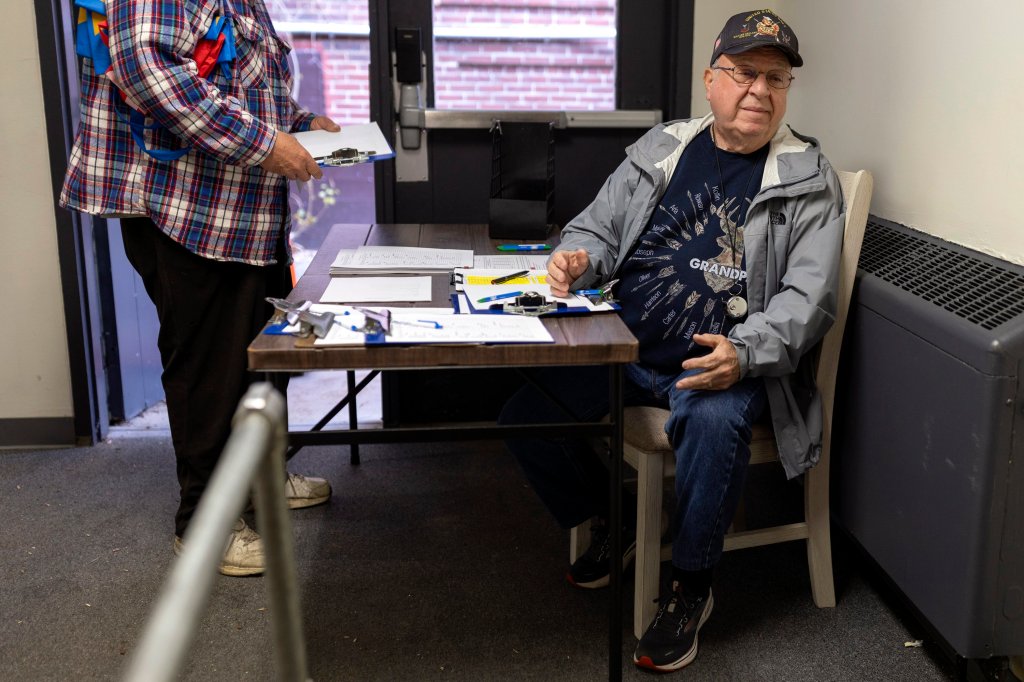A man sits at a table with clipboards as another person holds a clipboard in front of him.