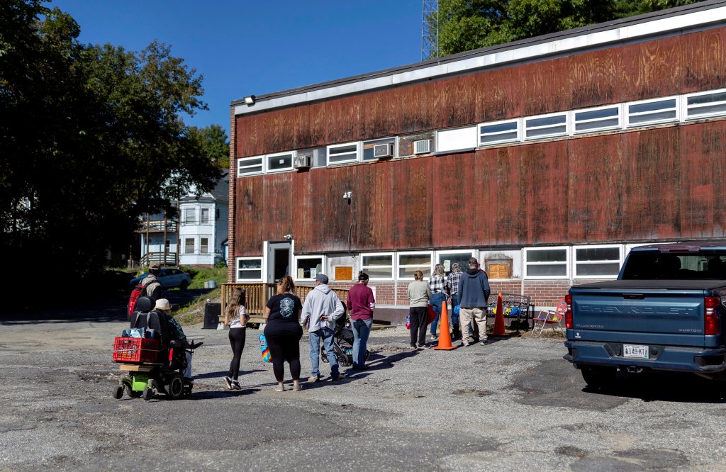People stand in line outside of an old, rusty looking building.