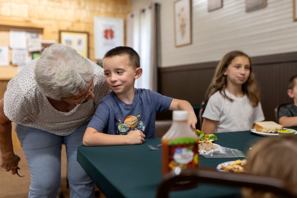 A young boy sitting at a table leans in to look at an older woman who's leaning down to his eye level.